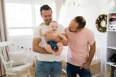 An LGBTQ+ couple holding their young toddler, who’s smiling and laughing